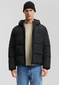 Chaqueta acolchada negra con cremallera frontal, capucha y dos bolsillos laterales. Presenta un diseño acolchado y puños elásticos. Se lleva con una camisa beige clara.