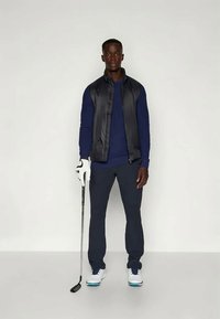 Čierny bezrukávový sveter cez modrú dlhú košeľu, spárovaný s námorníckymi nohavicami a bielymi golfovými topánkami. Drží golfovú palicu s bielou rukavicou.