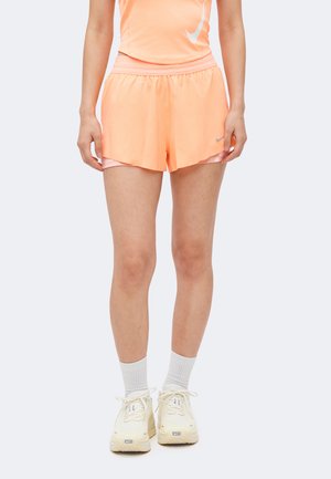 Osoba ubrana w brzoskwiniowe, warstwowe szorty sportowe, pasujący top, białe skarpetki i białe buty do biegania Nike, stoi prosto.