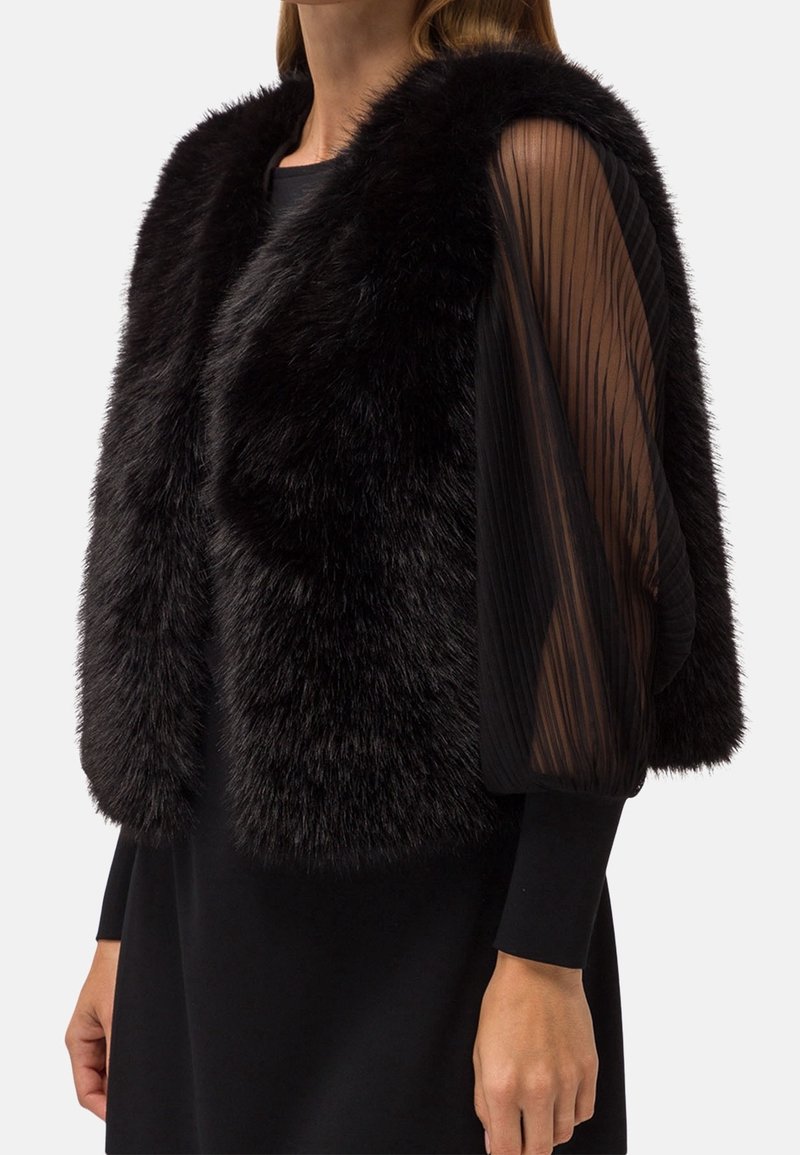 Svart faux fur väst med en plyschliknande textur, med en rundad krage och genomskinliga, plisserade svarta ärmar för kontrast och elegant lager på lager.