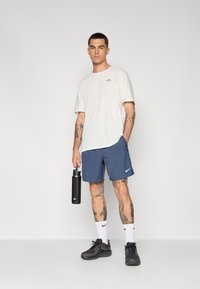 Men's athletic outfit featuring a cream t-shirt, blue shorts, black sneakers, and white socks with a logo, holding a black water bottle.