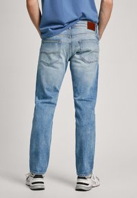 Hombre vistiendo unos vaqueros rectos de color azul claro, zapatillas blancas y una camisa azul, de pie con las manos en los bolsillos mirando hacia atrás.