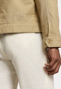 Detailní záběr ruky osoby blízko kyčle, která má na sobě béžovou bundu a světlé krémové texturované kalhoty s jemnou vlhkou skvrnou.