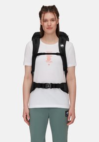 Czarny plecak z wieloma regulowanymi paskami, noszony na białej koszuli z pomarańczowym grafiką. Model stoi na tle jednolitego tła.