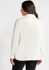 Biała koszula z długim rękawem i kołnierzykiem, o gładkiej fakturze i falbaniastych mankietach, w zestawieniu z czarnymi spodniami. Widok z tyłu, bez wzorów ani akcentów.