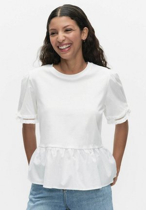 Vrouw glimlachend, draagt een witte blouse met korte mouwen en ruches aan de zoom, en mouwen met kantafwerking, gecombineerd met een blauwe spijkerbroek, tegen een witte achtergrond.