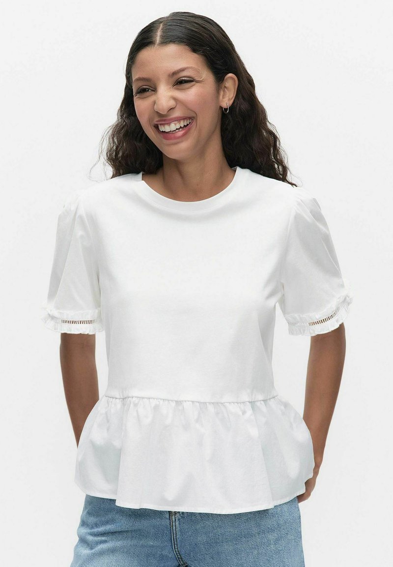 Kvinde smilende iført hvid kortærmet bluse med flæsekant og ærmer med blondekant, kombineret med blå jeans, foran hvid baggrund.