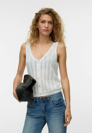 Kobieta z kręconymi włosami w białym, bezrękawnym dzianinowym topie i niebieskich dżinsach trzyma czarną skórzaną kopertówkę na jednolitym tle.