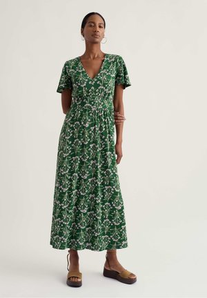 Nő zöld virágos V-nyakú midi ruhában, rövid ujjúval és barna platform szandálban, sima világos háttér előtt állva.