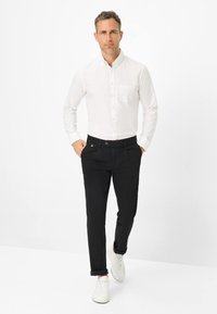 Weißes Hemd mit Knopfleiste und Brusttasche, schwarze Hose und weiße Sneakers. Glattes Material, taillierte Passform, mit hochgerollten Hosenbeinen.