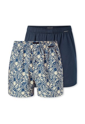 Shirtless man with short hair wearing blue and beige patterned boxer shorts, standing with arms crossed against a plain background.