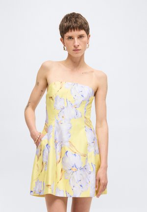 Femme aux cheveux bruns courts portant une robe jaune bustier avec un grand imprimé floral blanc et bleu, debout devant un fond uni.