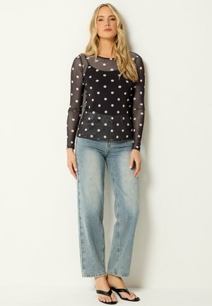 Kvinde med langt blondt hår iført en sort transparent top med lange ærmer og polkaprikker, lyseblå højtaljede jeans og sorte sandaler.