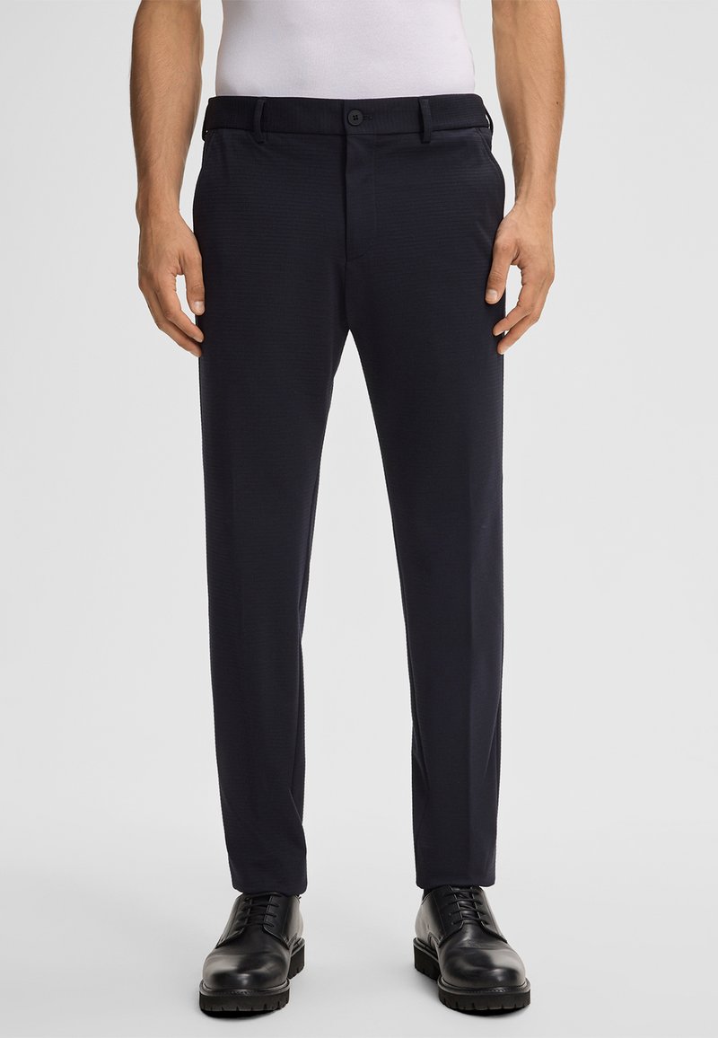 Pantaloni tailored blu navy realizzati in un tessuto liscio e testurizzato, con tasche frontali e passanti per la cintura; abbinati a scarpe nere con lacci.