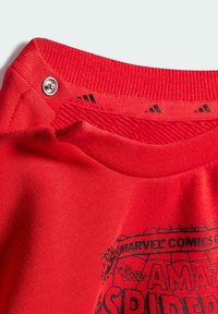 T-shirt en coton rouge avec un col côtelé et un bouton pression. L'avant présente un texte graphique noir et un design de toile d'araignée.