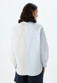 Button-up Shirt mit einem blauen und weißen gestreiften Design auf einer Seite und einfarbig weiß auf der anderen Seite. Lange Ärmel, entspannte Passform.