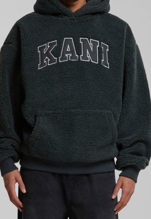 Person trägt einen schwarzen, flauschigen Hoodie mit Fronttasche und großen weißen Umrissbuchstaben „KANI“ auf der Brust, kombiniert mit schwarzen Hosen.