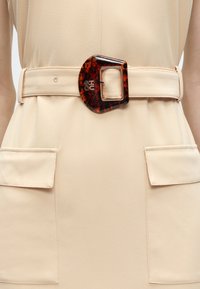 Μπεζ φόρεμα με μεγάλες μπροστινές τσέπες και φαρδιά μπεζ ζώνη με οβάλ αγκράφα από χελώνα με μεταλλικό καρφί.