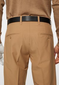 Bruna byxor med en slät tygfinish, utrustad med två bakfickor och ett svart perforerat bälte. Hand i en vit handske synlig.