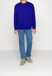 Persona con un maglione blu reale semplice, jeans azzurri e sneakers grigie, in piedi contro uno sfondo chiaro.