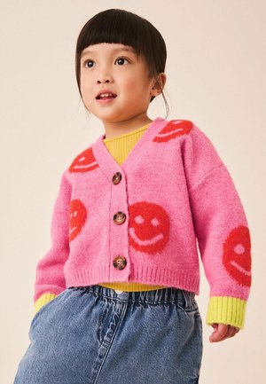Copil mic care poartă un cardigan roz cu fețe zâmbitoare roșii, cămașă galbenă cu mâneci lungi și blugi albaștri, uitându-se ușor într-o parte.