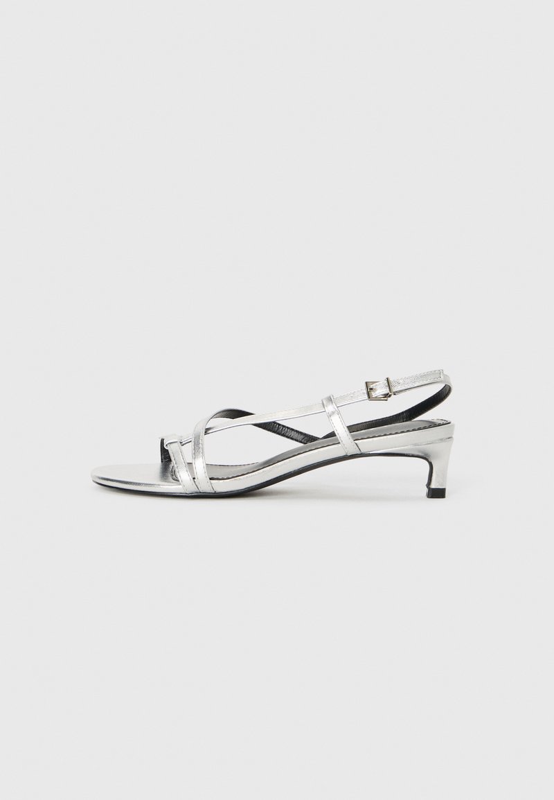 Sandaletti slingback metallici argento con tacco basso, caratterizzati da cinturini incrociati e una superficie liscia. Design minimalista.