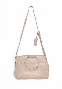 Béžová kožená kabelka s kulatým zlatým akcentem, nastavitelným ramenním popruhem a malým odnímatelným štítkem. Hladký povrch a obdélníkový tvar.