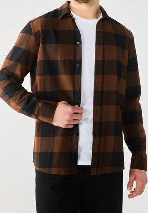 Homme boutonnant une chemise à carreaux marron et noir par-dessus un t-shirt blanc, portant un pantalon noir, sur un fond clair uni.