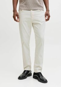 Pantaloni beige chiaro con vestibilità sagomata, dotati di tasche frontali e una texture liscia. Abbinati a scarpe eleganti nere per contrasto.