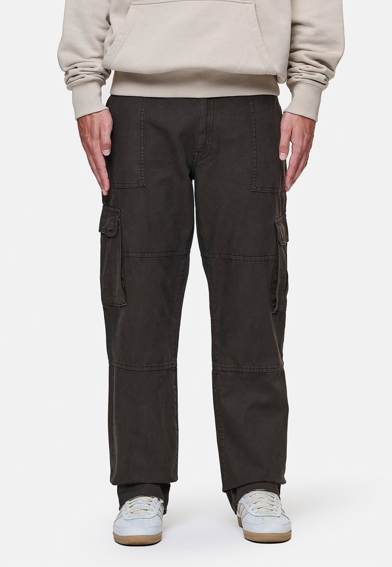 Personne portant un pantalon cargo noir et des baskets blanches, debout avec les mains détendues le long du corps devant un fond uni.