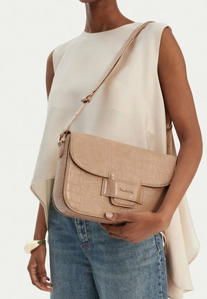 Donna con top beige senza maniche e jeans blu, che tiene una borsa a tracolla beige con texture e logo "GINO ROSSI" sulla patta frontale.