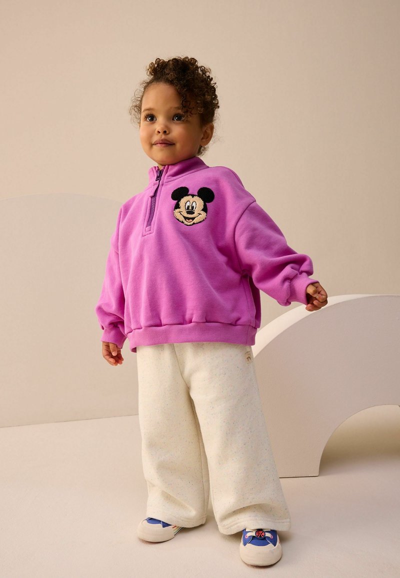 Lila Fleece-Sweatshirt mit Reißverschluss und gesticktem Mickey Mouse Design. Kombiniert mit cremefarbenen, weit geschnittenen Hosen und bunten Sneakern.