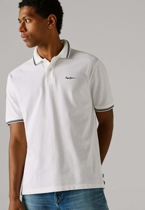 Bela polo majica s črnim izvezenim logotipom, ima klasičen ovratnik z kontrastnimi črtami na ovratniku in na rokavih. Oblečena z modrimi džinsi.