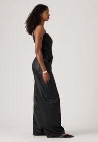 Svart denimkorsett med tunna axelband, kombinerad med vida svarta byxor med slät textur och subtil glans, sedd från sidan.