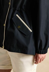 Tumši zils vējjakas mētelis ar elastīgām piedurknēm, apstrādāts ar baltu apdari un sānu kabatu, kurā ir misiņa pogcaurums.