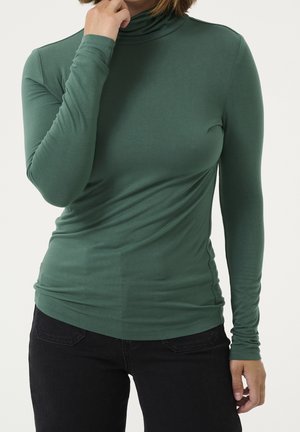 Femme portant un col roulé vert à manches longues et un pantalon noir, une main touchant son menton et l'autre détendue le long du corps.