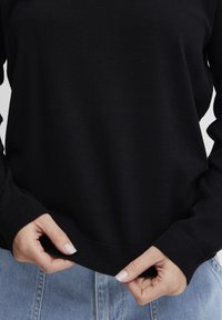 Črn pleten pulover z okroglim izrezom in rebrastimi manšetami. Tkanina se zdi mehka z gladko teksturo, kar prikazuje preprost in klasičen dizajn.