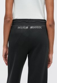 Černé tepláky Nike Shox s elastickým pasem a bílým vyšívaným logem na zadní straně. Měkká látka s hladkou texturou.