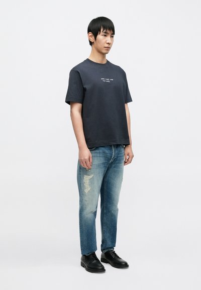 Μπλε ναυτικό βαμβακερό t-shirt με λευκό κείμενο, συνδυασμένο με ανοιχτό μπλε σκισμένο τζιν και μαύρα παπούτσια με κορδόνια, στέκεται σε απλό φόντο.