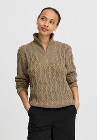 Gebreide trui in taupe met een hoge kraag en rits, voorzien van een gestructureerd diamantenpatroon en losse mouwen, gecombineerd met zwarte pantalon.