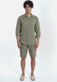 Olijfgroene linnen shirt met opgerolde mouwen, bijpassende shorts en beige gestreepte instappers. Eenvoudig ontwerp, relaxte pasvorm en minimale details.