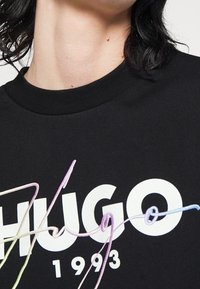 Zwart katoenen T-shirt met gedurfde witte "HUGO" tekst, geaccentueerd met meerkleurige borduursels in gebogen lijnen. Opvallende ronde halslijn.