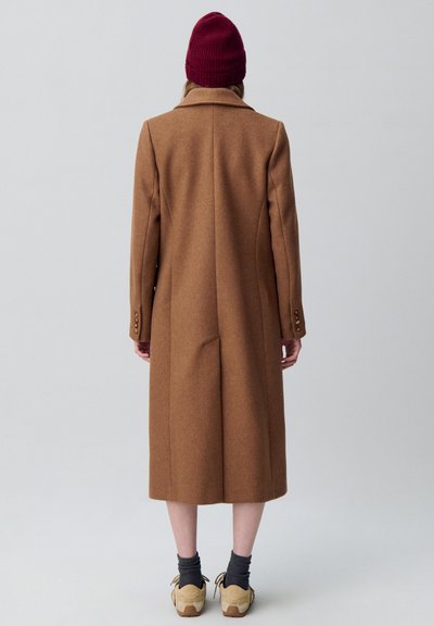 Cappotto di lana marrone con un design strutturato, lunghezza lunga e revers a punta; indossato con un berretto di lana rosso e scarpe beige.