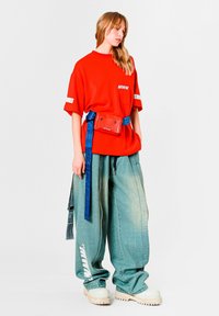 Camiseta roja de gran tamaño con logo, cinturón azul y pantalones anchos de mezclilla azul claro. Zapatos plataforma gruesos color crema. Fondo blanco.