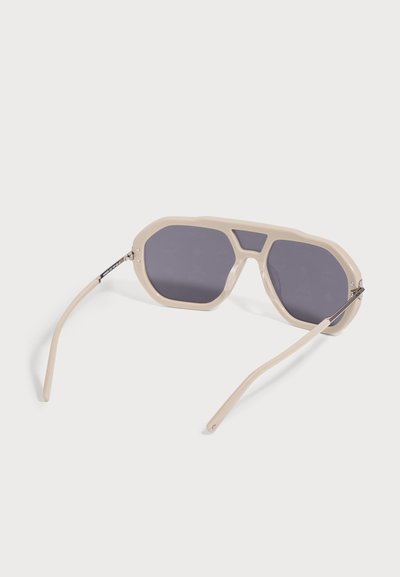 Lunettes de soleil oversize beige avec verres gris foncé et branches fines posées à plat sur un fond blanc.