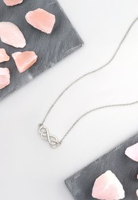 Sølv uendelighedsnære med glitrende accenter på en delikat kæde, sat mod grå skifer og rosa saltkrystaller.
