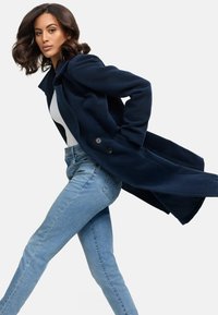 Navyblauwe lange jas met een op maat gemaakt ontwerp, voorzien van twee knopen en een vloeiende onderkant. Draag het over een witte top en lichtblauwe skinny jeans.
