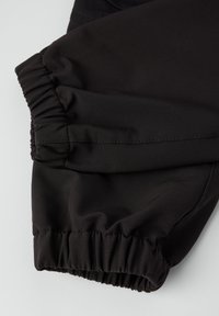 Close-up van zwarte stoffen broekspijpen met elastische boorden, die de gegarneerde en gestikte details op een lichte achtergrond benadrukken.