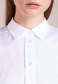 Camicia bianca con colletto, dalla texture liscia, con una pattina di bottoni e bottoni trasparenti sul davanti, che mostrano linee nette e pulite.