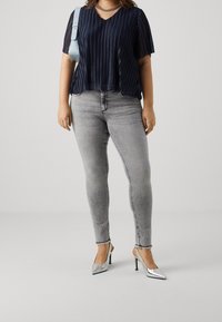 Kvinna som bär en genomskinlig marinblå plisserad blus, ljusgrå smala jeans med fransade kanter, silverfärgade spetsiga klackar, kedjehalsband och en ljusblå axelväska.